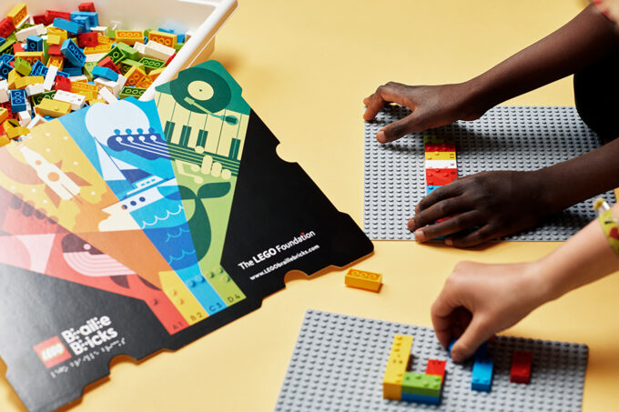 Legoklossar, legopaket och två par barnhänder som monterar Lego Braille Bricks legon på underlag, vilka utvecklats för att stödja inlärning av punktskrift.