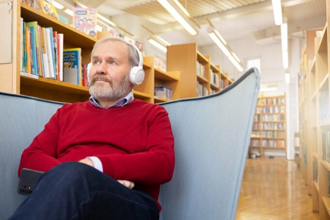 Henkilö istuu kirjastossa nojatuolissa ja kuuntelee äänikirjaa kuulokkeilla.