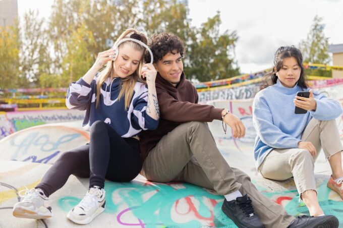 Kolme henkilöä istuu graffitein koristellussa skeittipuistossa. Yksi kuuntelee äänikirjaa kuulokkeilla, toinen nojaa häneen, kolmas katsoo puhelinta kädessään.