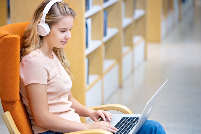 Henkilö istuu hymyillen kirjastossa nojatuolissa kannettava tietokone auki sylissään ja kuuntelee äänikirjaa valkoisilla kuulokkeilla.