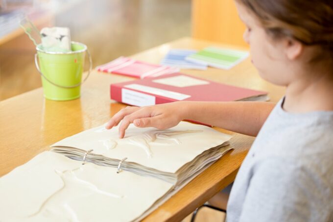 Ett barn sitter vid ett bord och känner på en reliefbild som föreställer en djur. Bilden finns i en mapp på bordet. På bordet finns ytterligare en mapp och skolmaterial.