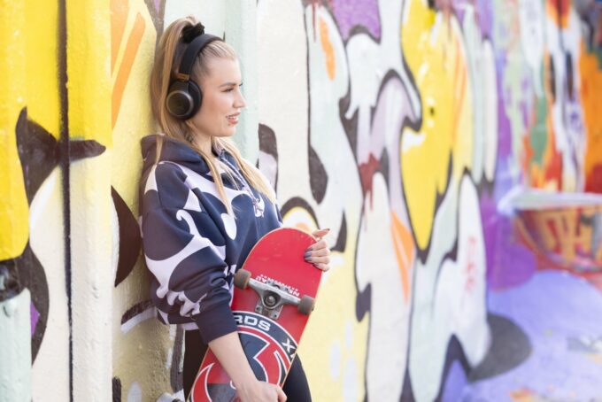 En person lutar sig mot en färgglad graffitivägg. Hen har en skateboard i handen och lyssnar på en talbok från Tillgänglighetsbiblioteket Celia med hörlurar.