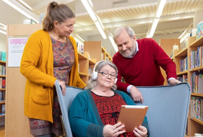 Tre personer på ett bibliotek. En av dem sitter i en fåtölj och lyssnar på en bok från Tillgänglighetsbiblioteket Celia med hörlurar. De andra två står bakom stolen.