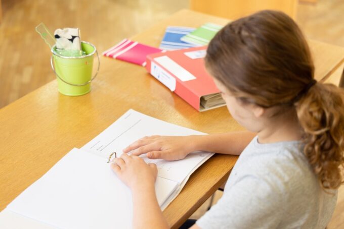 Ett barn, bakifrån, som läser en punktskriftsbok vid bordet. På bordet finns förutom boken en röd pärm, färgglada anteckningsböcker och kontorsmaterial i en grön kopp.