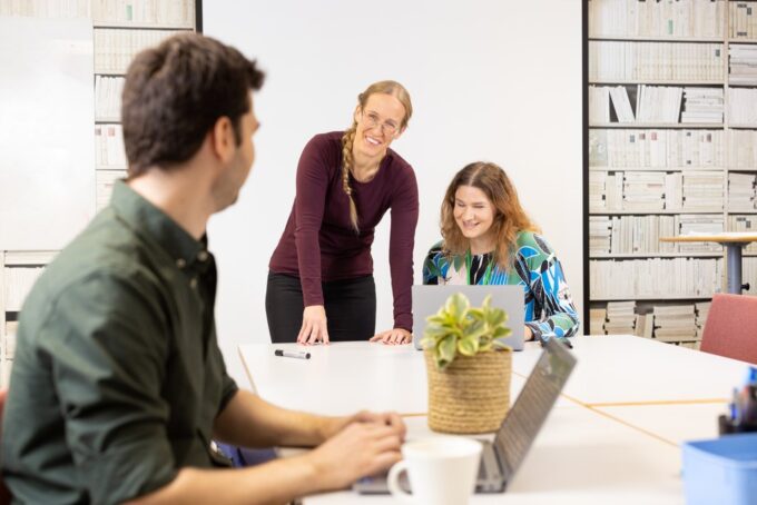 Tre personer i ett konferensrum. Två sitter, en står. På bordet står bärbara datorer och en växt. Alla ler.