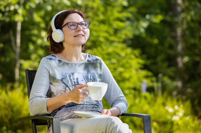 En person sitter ute i trädgården med en kaffekopp i handen och lyssnar på en talbok med hörlurar.